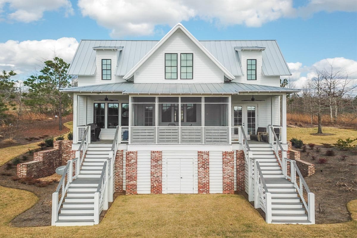 House plan exterior. Two-story farmhouse with metal roof, screened porch, dormers, and brick foundation with staircases on either side.