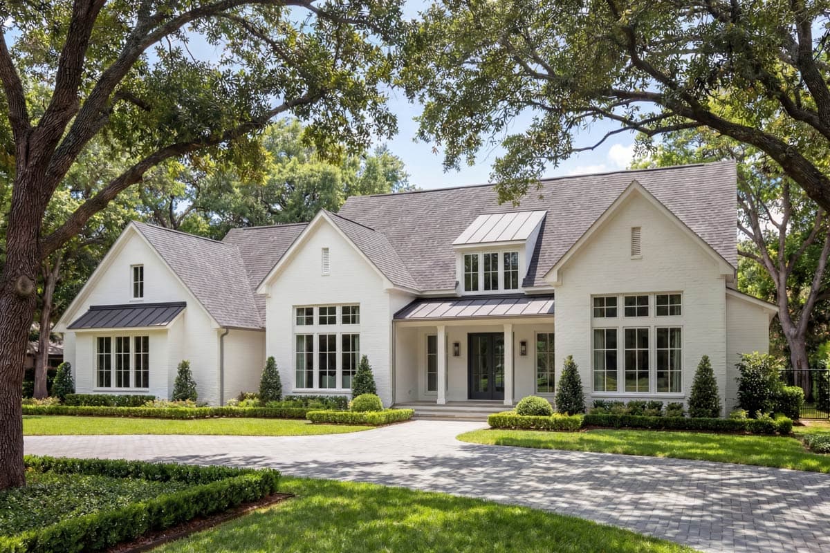 White brick home with dark roof and trim, under lush tree canopy. A circular paved driveway leads to the front door, surrounded by greenery.