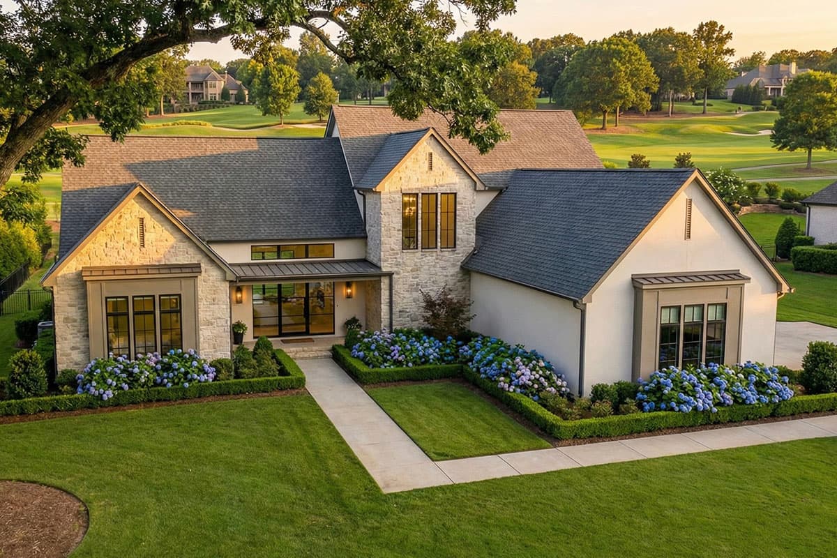 Exterior view of a modern house with stone and white facade, large windows, and a manicured lawn. Lush trees in the background.