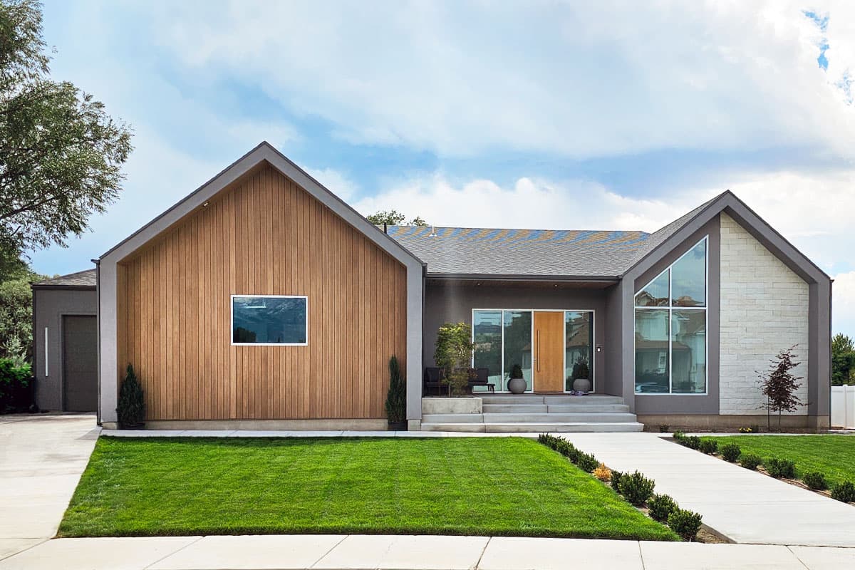 House plan exterior. Modern style, one story. Features include wood siding, multiple gables, large windows, and a covered entry with a wooden door.
