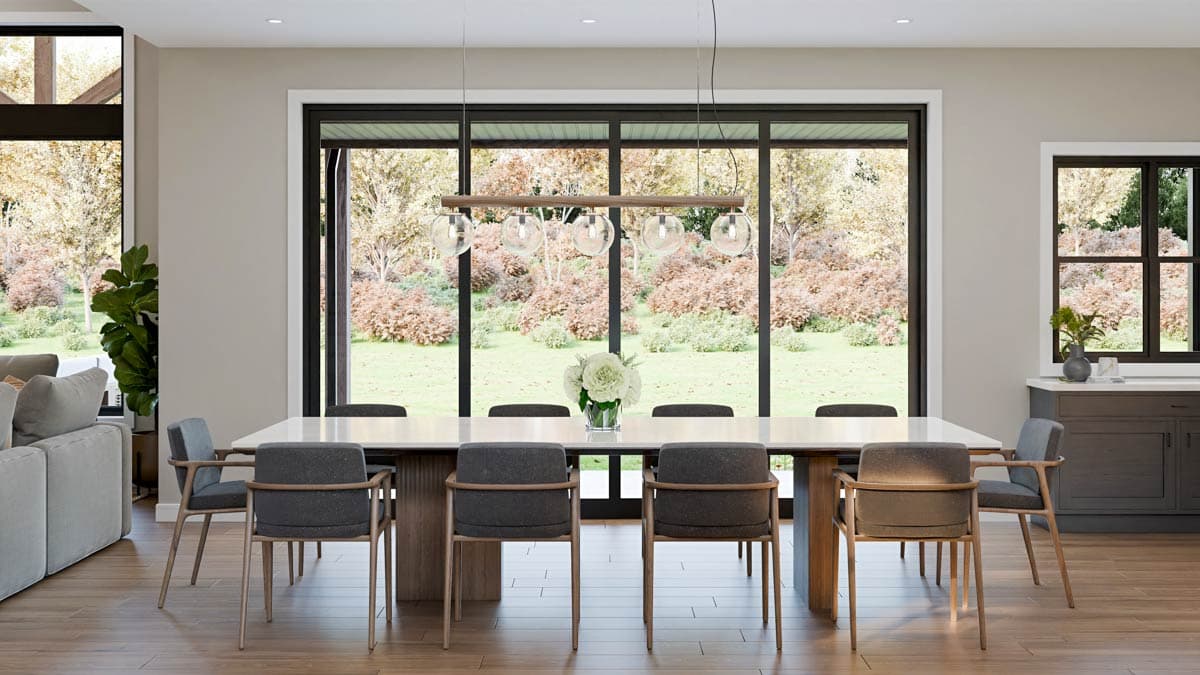 Modern dining room with a long white table, eight chairs, and large windows overlooking a backyard with trees and flowers.